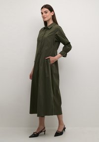 Olive green button-up dress with long sleeves, elastic cuffs, and side pockets. Designed with a knee-length A-line silhouette, smooth texture.