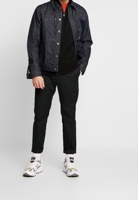 Chaqueta de denim en azul oscuro con cierre de botones, superpuesta a una camiseta negra. Combinada con pantalones negros ajustados y zapatillas blancas con detalles.