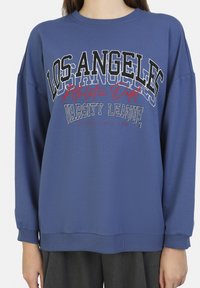 Sudadera azul con gráficos de "LOS ÁNGELES Athletic Dept. VARSITY LEAGUE" en blanco, negro y rojo. Corte relajado con puños acanalados.