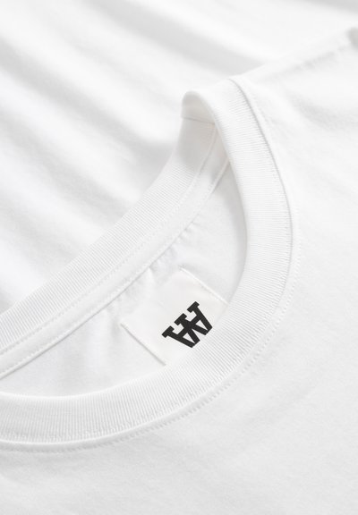 Κοντινό πλάνο λευκού T-shirt με στρογγυλή λαιμόκοψη που δείχνει τις ραφές του ριμπ λαιμού και μια ασπρόμαυρη ετικέτα μάρκας μέσα στη λαιμόκοψη.