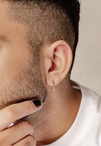 Silberner Creolenohrring an einem kurz geschorenen Ohr mit kurzem, strukturiertem Haar. Die Person hat eine Hand nahe ihres Kinns, mit schwarz lackierten Fingernägeln.