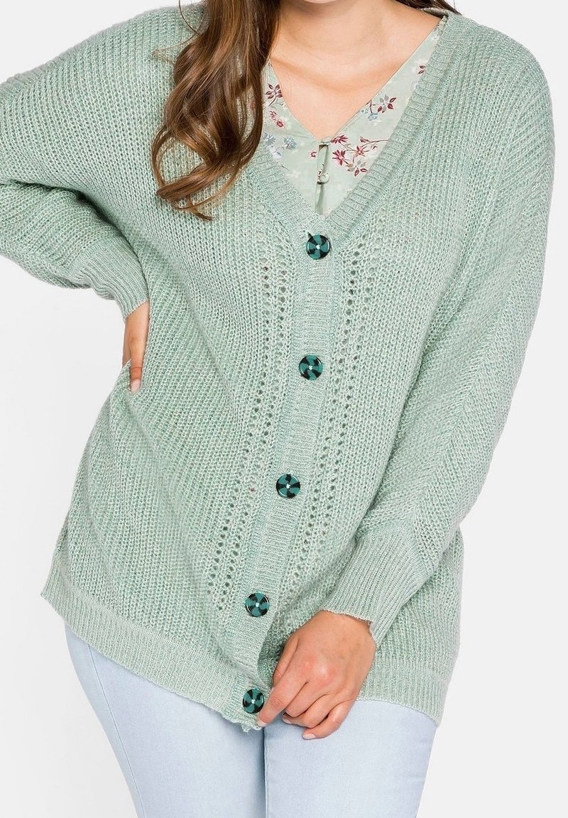 Femme portant un cardigan tricoté vert clair avec de gros boutons verts sur un haut à motif floral, associé à un jean bleu clair.