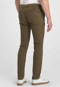 Man in slim-fit olijfgroene broek en witte sneakers, staande met de rug naar de camera tegen een effen achtergrond.
