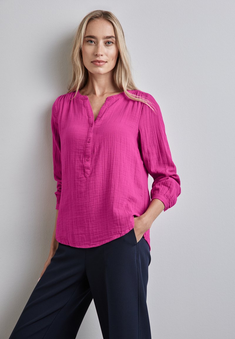 Blusa lunga a maniche lunghe di colore rosa, con texture, colletto a scollatura a V e patta con bottoni, abbinata a pantaloni scuri. Il modello posa con una mano in tasca.
