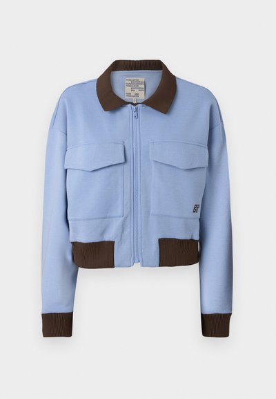Lyseblå cropped jakke med en brun ribbeformet krage og mansjetter, frontlås med glidelås og to brystlommer. Glatt stoff med en uformell design.