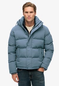 Lyseblå puffjakke med hætte, prydet med et quiltet design, forkert snaplukning og ribbede manchetter, båret over en mørk sweater.