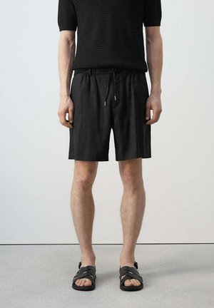 Hombre con camisa negra de manga corta texturizada, pantalones cortos negros con cordón y sandalias negras, de pie frente a un fondo claro y liso.