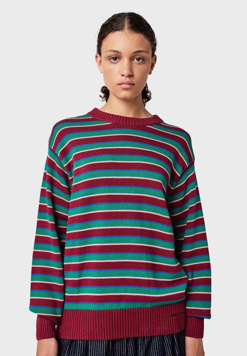 Giovane donna con capelli ricci scuri che indossa un maglione lavorato a maglia a maniche lunghe, largo, con strisce orizzontali di bordeaux, verde, blu e bianco.