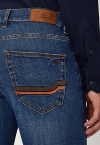 Jeans denimowe w ciemnoniebieskim kolorze, z brązową skórzaną naszywką i kontrastowym brązowo-pomarańczowym przeszyciem na tylnej kieszeni.