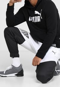 Sudadera negra con el logo blanco de PUMA; combinada con pantalones de chándal que tienen paneles en blanco y negro. Zapatillas deportivas grises con detalles blancos.