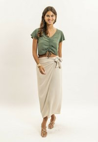 Haut court vert avec un détail de fronces à l'avant, associé à une jupe portefeuille beige avec un nœud à la taille et une texture douce. Sandales beiges à talons.