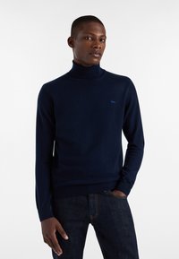 Mornarsko moder pulover z visokim ovratnikom, izdelan iz mehkega materiala, z majhnim svetlomodrim logotipom na prsih ter rebrastimi manšetami in robom.