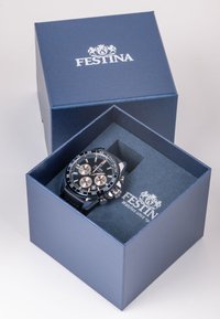 Festina pulkstenis zilā dāvanā kastē. Pulkstenim ir melns ciparnīca, sudraba apdare un melna siksna, novietots uz mīksta auduma oderes.