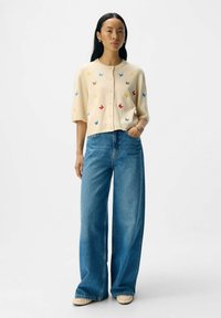 Gilet crème à manches courtes avec des broderies de papillons colorés, associé à un jean bleu à jambes larges et des chaussures de couleur claire.