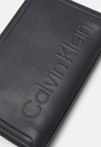 Svart läderplånbok med präglad "Calvin Klein"-logotyp. Har en slät textur, syddetaljer och en rektangulär form.