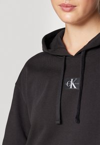 Felpa nera con cappuccio realizzata in tessuto morbido, con cappuccio regolabile tramite stringhe e un'etichetta quadrata con il testo "Calvin Klein Jeans".