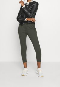 Mustat nahkatakki, valkoinen pyöreäkauluksinen paita, oliivinvihreät joggerit kavennetuilla lahkeilla ja valkoiset lenkkarijalkineet vaaleilla yksityiskohdilla.