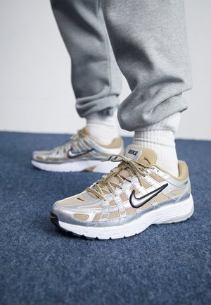 Osoba nosząca beżowo-srebrne buty sportowe Nike, białe skarpetki i szare spodnie dresowe stoi na niebieskim dywanie.