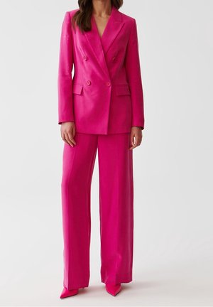 Woman wearing bright pink double-breasted blazer with matching wide-leg trousers and pointed shoes, standing against plain gray background.