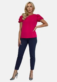 Fuksjowa bluzka z falbanowymi rękawami, zestawiona z granatowymi spodniami w kropki. Modelka w srebrnych szpilkach, stojąca w neutralnej pozycji.