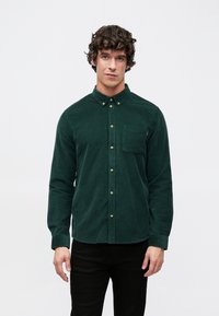 Camicia verde in velluto a coste con colletto abbottonato, tasca frontale, bottoni dorati e maniche lunghe, abbinata a pantaloni neri.