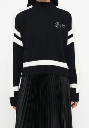 Femme portant un pull noir à rayures blanches avec le logo "Guess Ski", associé à une jupe plissée noire.