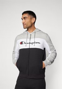 Grå, vit och svart zip-up hoodie med snoddar och "Champion"-logotyp. Har en färgblockerande design och sidofickor. Mjuk tygtextur.