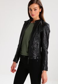 Black leather jacket with textured shoulder accents, zippered pockets, and a fitted design, paired with a green top and black pants.