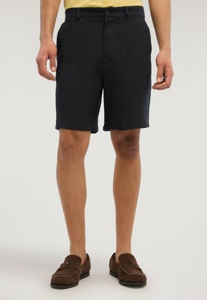 Mand iført sorte skræddersyede shorts og brune ruskindslædermokassiner stående mod en ensfarvet baggrund.