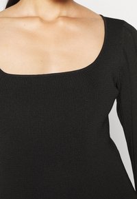 Donna che indossa un top a costine nero con una ampia scollatura quadrata e maniche lunghe, mostrata dalle spalle alla vita.