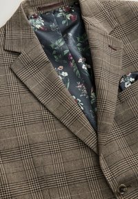 Braun karierter Blazer mit strukturiertem Außenmaterial und floral gemusterter Satin-Fütterung. Bemerkenswerte Details sind ein Brusttaschentuch.