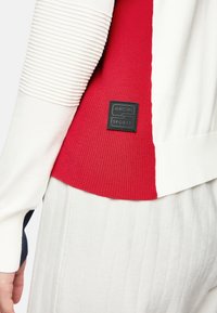 Rot-weißer, gerippter Sportsweater mit strukturierten Ärmeln. Verfügt über ein schwarzes Logo-Patch im unteren Rückenbereich. Der Stoff wirkt weich und elastisch.