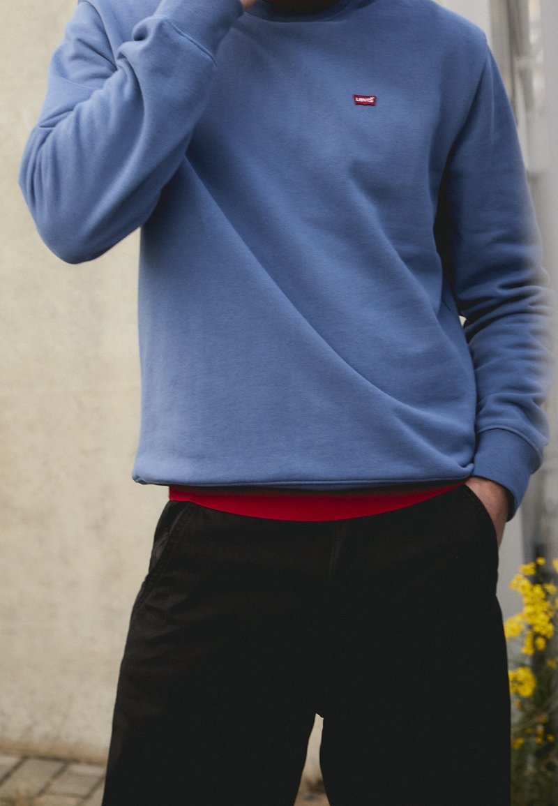 Sudadera azul claro con cuello redondo y pequeño logo rojo en el pecho, superpuesta sobre una camisa roja, combinada con pantalones negros.