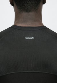 Melna sporta krekls ar fakturētu audumu, ar plakaniem šuvumiem un mazu VIGOR7 logotipu aizmugurē pie kakla līnijas.