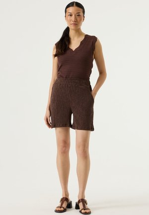 Braunes ärmelloses Oberteil mit einer gezackten Ausschnittkante, kombiniert mit braunen strukturierten Shorts mit elastischem Bund und Taschen. Braune Sandalen mit Absatz.