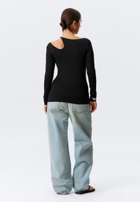 Schwarzes Langarm-Oberteil mit einem Ausschnitt am Schulterbereich, kombiniert mit hellblauen, locker sitzenden Denim-Jeans. Glattes Material, figurbetonter Schnitt.