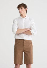 Camicia bianca leggera con bottoni e pantaloni corti marrone testurizzati, dotata di chiusura a bottoni e due tasche laterali, design lungo fino al ginocchio.