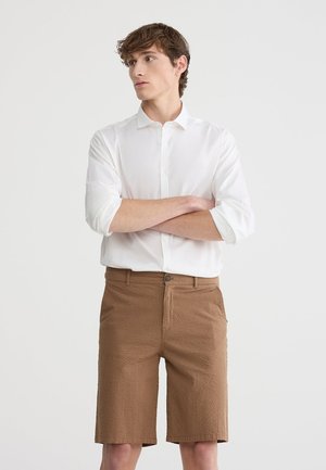 Camicia bianca leggera con bottoni e pantaloni corti marrone testurizzati, dotata di chiusura a bottoni e due tasche laterali, design lungo fino al ginocchio.
