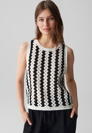 Jeune femme aux cheveux châtain clair porte un haut en crochet blanc sans manches avec des rayures verticales noires et un pantalon noir, mains dans les poches.