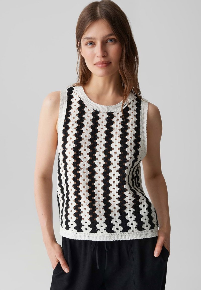 Jeune femme aux cheveux châtain clair porte un haut en crochet blanc sans manches avec des rayures verticales noires et un pantalon noir, mains dans les poches.