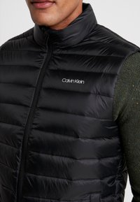 Czarna kamizelka puchowa z wysokim kołnierzem, z poziomym pikowaniem i przednim zamkiem błyskawicznym. Logo "Calvin Klein" widoczne na piersi.