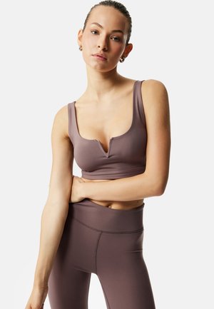 Joven con el cabello recogido, vistiendo un sujetador deportivo malva y leggings a juego de cintura alta, de pie con un brazo cruzado sobre la cintura.