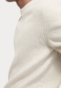 Stickad tröja i off-white, med ett texturerat mönster och rund halsringning. Tyget verkar mjukt och något andningsbart.