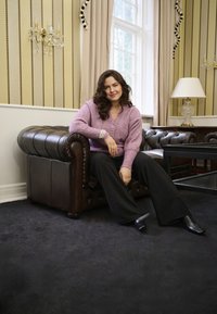 Gebreide roze trui met een V-hals, gecombineerd met donkere wijde broek. Zittend op een bruine leren tufted bank tegen gestreepte muren.