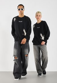 Black sweatshirts with crane prints, crew necklines, and ribbed cuffs, paired with distressed gray wide-leg jeans and sneakers.