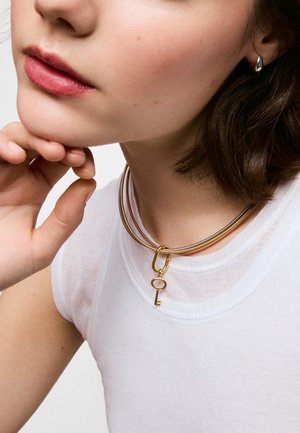 Primer plano de una persona que lleva una camiseta blanca sin mangas, un collar tipo choker dorado con un colgante de llave y un pendiente de aro plateado.