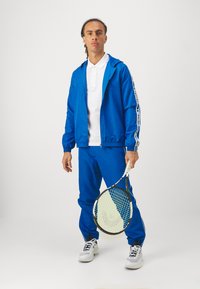 Kék melegítő szett zipzáras dzsekivel és rugalmas mandzsettákkal, fehér pólóinggel párosítva. Teniszütőt tart a kezében, fehér sportcipőt visel.