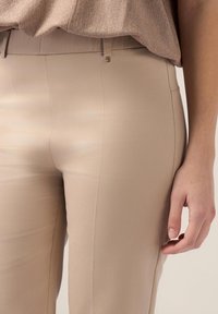 Beige op maat gemaakte broek met een gladde textuur en smalle pasvorm. Voorzien van een sluiting aan de voorkant en zijzakken. Weergegeven met een hand die de stof vasthoudt.