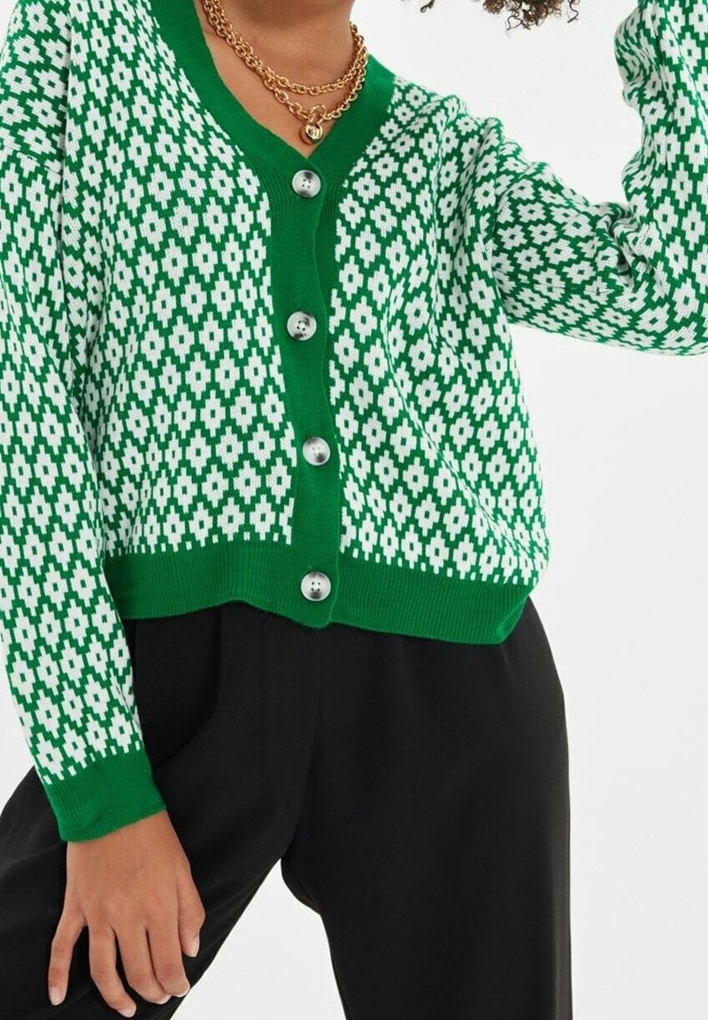 Cardigan boutonné à motifs verts et blancs porté avec un pantalon noir, accessoirisé de colliers à chaînes dorées.