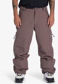 Pantalons neige légers, mauves, avec une coupe ample, plusieurs poches zippées et des poignets ajustables, associés à des baskets noir et blanc.